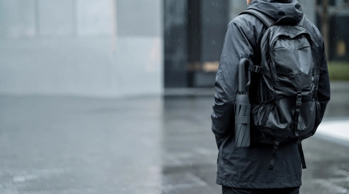 Personne vue de dos sous la pluie, un parapluie Nuvya StormHook plié accroché à son sac à dos par le mousqueton